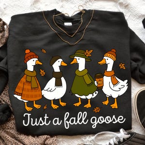 May include: Dark gray sweatshirt featuring a whimsical illustration of four geese dressed in fall attire. The geese wear hats, scarves, and coats, with one holding a coffee cup. The text "Just a fall goose" is printed below the geese.