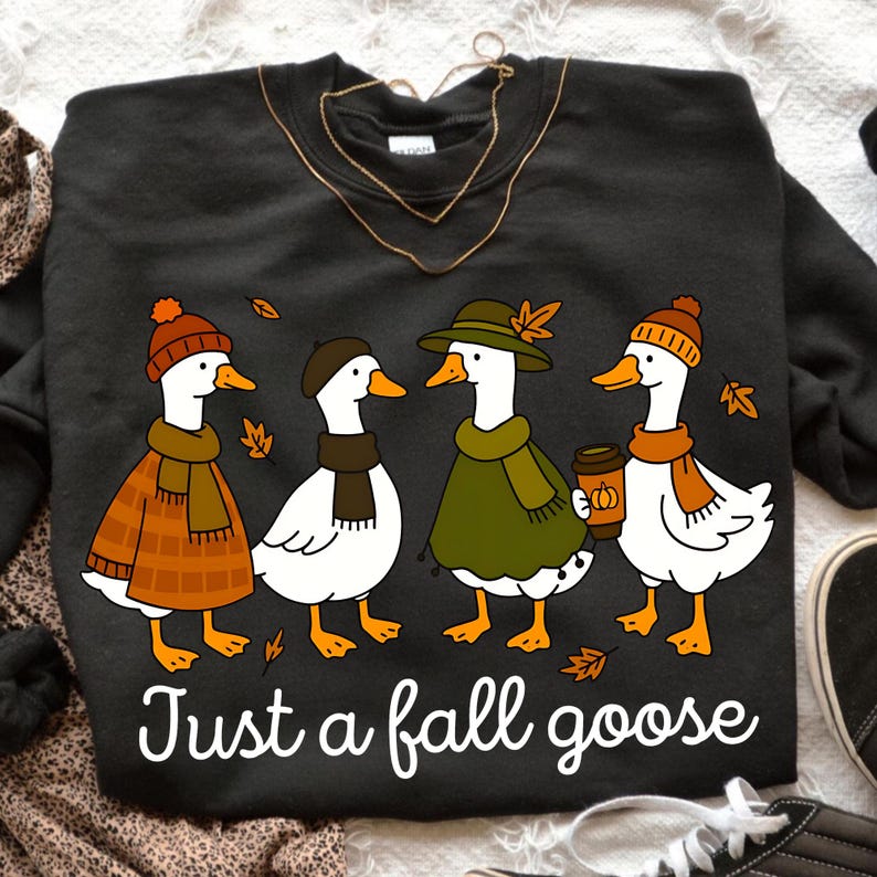 May include: Dark gray sweatshirt featuring a whimsical illustration of four geese dressed in fall attire. The geese wear hats, scarves, and coats, with one holding a coffee cup. The text "Just a fall goose" is printed below the geese.