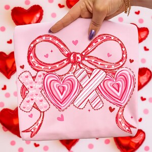 May include: A pale pink t-shirt with a large red bow and the letters "XOX" in a heart and candy design. The shirt has a Valentine's Day theme, with small hearts scattered around the design.