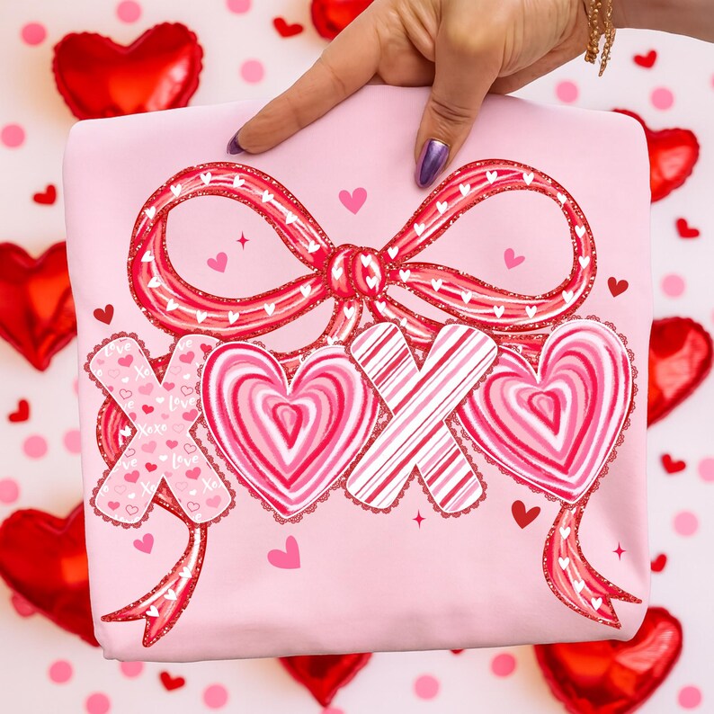 May include: A pale pink t-shirt with a large red bow and the letters "XOX" in a heart and candy design. The shirt has a Valentine's Day theme, with small hearts scattered around the design.
