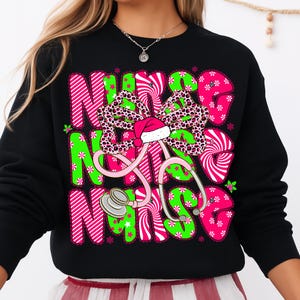 May include: Black sweatshirt with a festive design. The design features the word "NURSE" in pink and green, a leopard print bow with a Santa hat, and a pink stethoscope. The design is decorated with snowflakes.