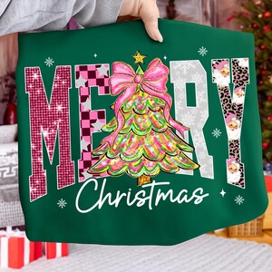 May include: A green Christmas-themed sweatshirt with the words "MERRY Christmas" in a festive design. The "M" is pink with glitter, the "E" is checkered, the "R" is white with snowflakes, and the "Y" has Santa faces. A colorful Christmas tree with a pink bow is in the center.