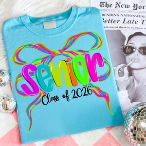 May include: A light blue t-shirt featuring the word "Senior" in vibrant, bubble letters, and the text "Class of 2026". The design incorporates a rainbow-colored bow and streamers. The shirt is displayed on a pink and white checkered surface.