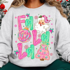 May include: A light gray sweatshirt with a colorful Christmas graphic. The graphic includes the words "Fald Lald" in pink and green bubble letters, a Santa Claus illustration, and a Christmas tree design.
