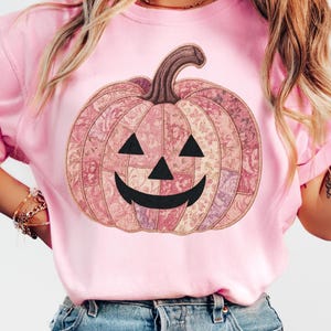 K&ouml;nnte beinhalten: Hellrosa Sweatshirt mit einem dekorativen K&uuml;rbisdesign. Der K&uuml;rbis hat ein Patchworkmuster in Rosa, Lila und Beige, mit schwarzen Dreiecksaugen und einem l&auml;chelnden Mund. Der Stiel ist braun.