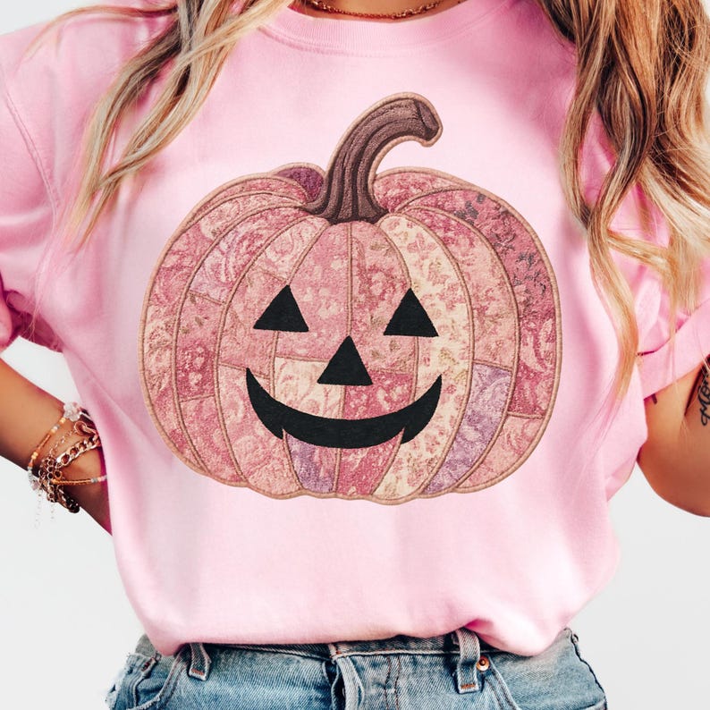 K&ouml;nnte beinhalten: Hellrosa Sweatshirt mit einem dekorativen K&uuml;rbisdesign. Der K&uuml;rbis hat ein Patchworkmuster in Rosa, Lila und Beige, mit schwarzen Dreiecksaugen und einem l&auml;chelnden Mund. Der Stiel ist braun.