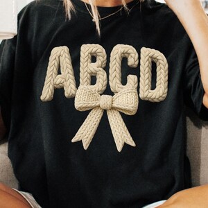 May include: Black t-shirt with the letters ABCD in a beige, knitted style, with a matching bow design. The letters are raised, creating a 3D effect. The overall design is simple and elegant.