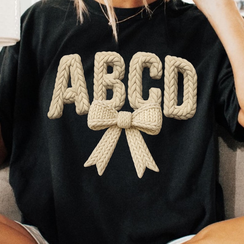 May include: Black t-shirt with the letters ABCD in a beige, knitted style, with a matching bow design. The letters are raised, creating a 3D effect. The overall design is simple and elegant.