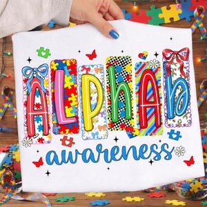May include: White t-shirt with the words "Alpha Awareness" in colourful, puzzle-piece-themed letters. The design includes butterflies, hearts, and bows, promoting autism awareness. The shirt is held by a person with blue nail polish.