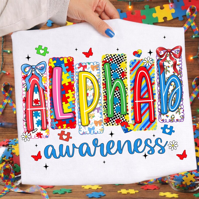 May include: White t-shirt with the words "Alpha Awareness" in colourful, puzzle-piece-themed letters. The design includes butterflies, hearts, and bows, promoting autism awareness. The shirt is held by a person with blue nail polish.