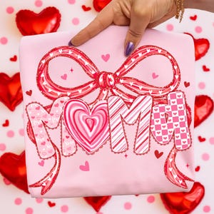 May include: A pink sweatshirt with a large red bow and the word "MOM" in a decorative font. The letters are adorned with hearts and patterns. Small hearts and stars are scattered around the design.