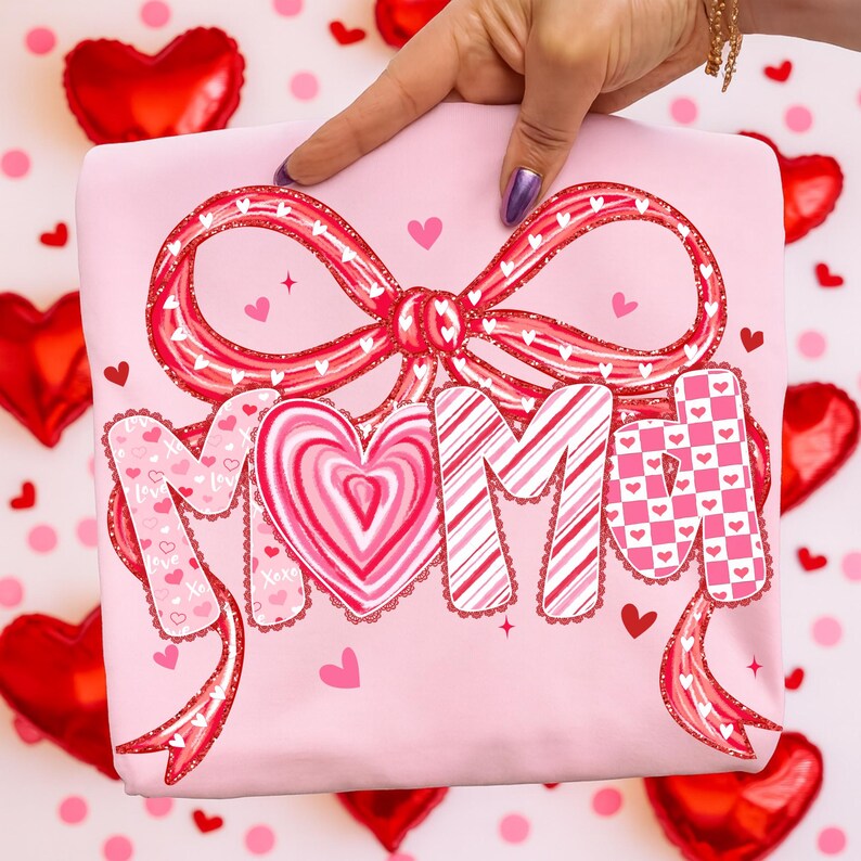 May include: A pink sweatshirt with a large red bow and the word "MOM" in a decorative font. The letters are adorned with hearts and patterns. Small hearts and stars are scattered around the design.