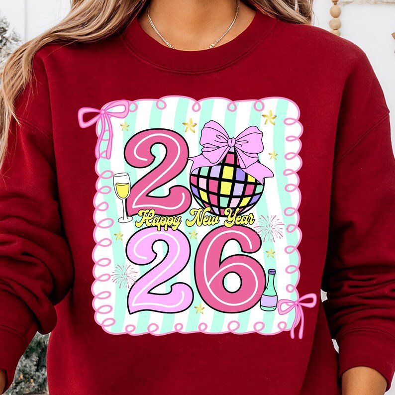 May include: Burgundy sweatshirt with a colorful graphic. The graphic features the numbers "2026" in pink, a disco ball, a champagne glass, and the words "Happy New Year". The design is set against a striped background.
