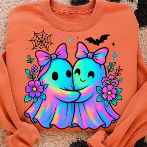 May include: Orange sweatshirt featuring a colorful graphic of two hugging ghosts with bows, surrounded by flowers, a spiderweb, and a bat. The ghosts have a rainbow-like appearance. A cute and friendly design.