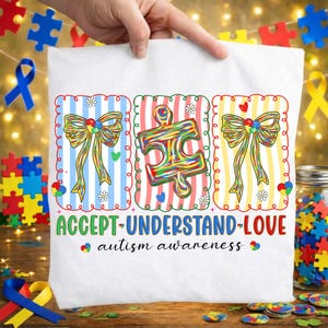 May include: White t-shirt with a colourful design promoting autism awareness. The design includes puzzle pieces, bows, and the words "ACCEPT-UNDERSTAND-LOVE." The shirt is held up by a hand, with puzzle pieces and ribbons in the background.