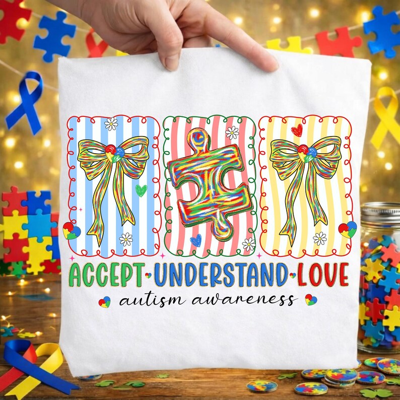 May include: White t-shirt with a colourful design promoting autism awareness. The design includes puzzle pieces, bows, and the words "ACCEPT-UNDERSTAND-LOVE." The shirt is held up by a hand, with puzzle pieces and ribbons in the background.