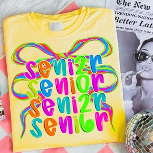 May include: A yellow t-shirt with the word "SENIOR" in colorful bubble letters. The word is repeated four times, with each instance in a different color. A rainbow ribbon design arches over the text.