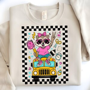 May include: Cream-colored sweatshirt featuring a cartoon raccoon wearing heart-shaped sunglasses and a pink shirt, sitting on a yellow school bus. The design includes a pink backpack, pencil, and school supplies, framed by a black and white checkered border.