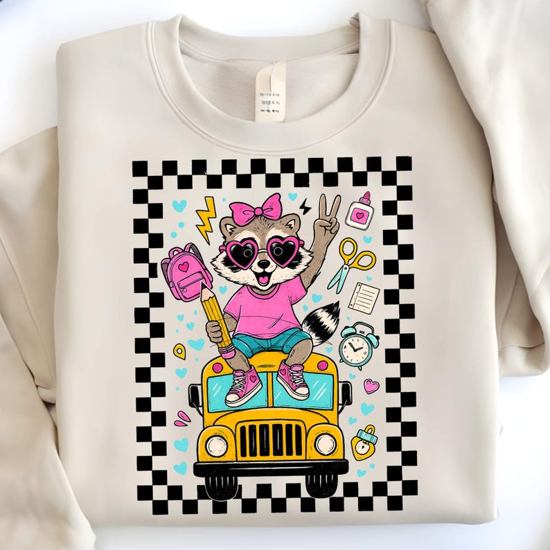 May include: Cream-colored sweatshirt featuring a cartoon raccoon wearing heart-shaped sunglasses and a pink shirt, sitting on a yellow school bus. The design includes a pink backpack, pencil, and school supplies, framed by a black and white checkered border.