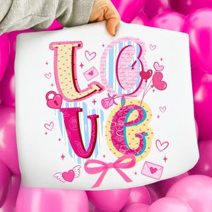 以下が含まれることがあります： 「LOVE」の文字が大きくカラフルに描かれた白い紙。文字はハート、バラ、リボンで装飾されています。背景はピンク色の風船のミックスです。