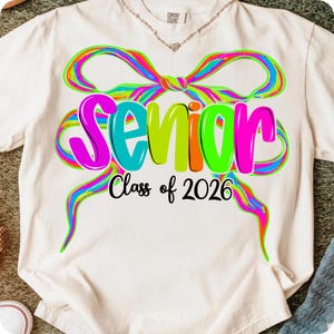 May include: Cream-coloured t-shirt featuring a colourful bow design and the word "Senior" in bright letters. Below, the text "Class of 2026" is printed in black. The shirt is a Comfort Colors brand.