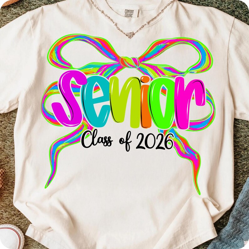 May include: Cream-coloured t-shirt featuring a colourful bow design and the word "Senior" in bright letters. Below, the text "Class of 2026" is printed in black. The shirt is a Comfort Colors brand.