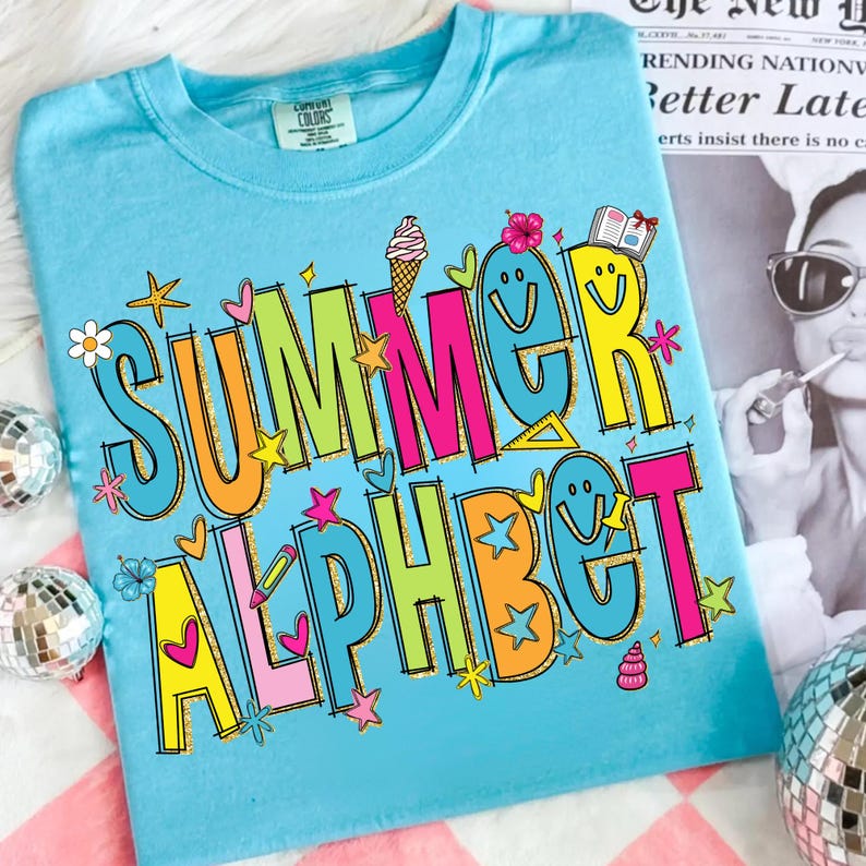 May include: A light blue t-shirt featuring the words "SUMMER ALPHABET" in vibrant, outlined letters. The design incorporates stars, hearts, flowers, and ice cream cones. The shirt is made of a soft material.