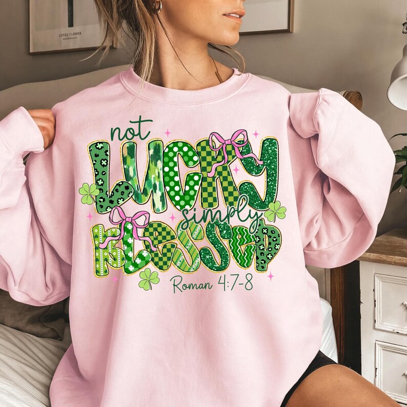 May include: A light pink sweatshirt with the text "not LUCKY simply BLESSED" in green, white, and pink. The design includes shamrocks and bows. The text "Roman 4:7-8" is printed below the main text.