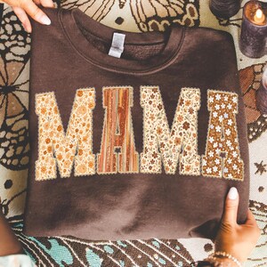 May include: A brown sweatshirt with the word "MAMA" in large, floral-patterned letters. The letters feature various designs, including flowers and stripes, in shades of orange, brown, and cream. The sweatshirt is held by hands.