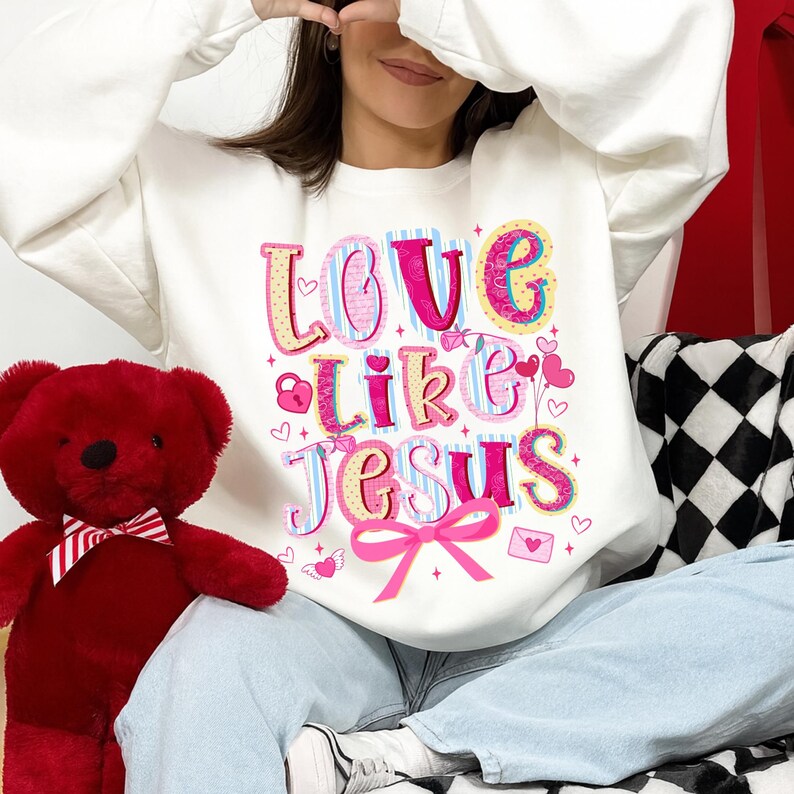 May include: White sweatshirt with the text "LOVE Like JESUS" in colorful, playful lettering. The design includes hearts, a bow, and other decorative elements. A red teddy bear with a bow tie is on the left.