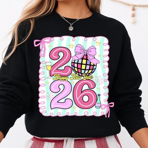May include: Black sweatshirt with a colorful graphic design for the New Year. The design includes the numbers "26" in pink, a disco ball, champagne, and the text "Happy New Year". The design is framed with a pink border and bows.