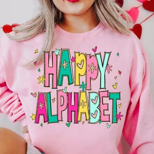 K&ouml;nnte beinhalten: Ein hellrosa Sweatshirt mit den Worten "HAPPY ALPHABET" in bunten, umrandeten Buchstaben. Die Buchstaben sind mit Sternen, Herzen und Blumen verziert. Das Sweatshirt hat einen Rundhalsausschnitt.