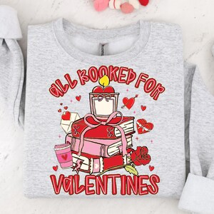 May include: A light gray sweatshirt with a graphic design featuring a stack of books, a candle, and hearts. The text "All Booked For Valentines" is in red. A rose and a coffee cup are also included in the design.