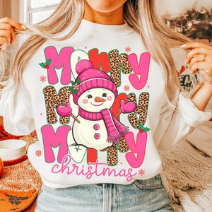 May include: White sweatshirt with a colorful Christmas design. The design features a snowman wearing a pink hat, scarf, and mittens, with the words "Merry Christmas" in pink and leopard print letters.