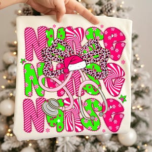 May include: A cream-colored t-shirt with a festive design. The words "NURSE" are displayed in pink and green, with a bow and Santa hat. A pink stethoscope is also featured. The design includes snowflakes and candy cane stripes.