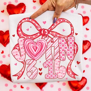 May include: White t-shirt featuring a large red bow with a heart-shaped design. The shirt has the text "JOIN 3 16" in a mix of pink and red patterns, including hearts and stripes. Small red hearts are scattered around the design.