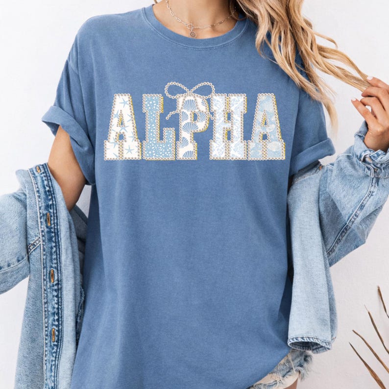 May include: A blue t-shirt featuring the word "ALPHA" in a decorative font. The letters are filled with a seashell and starfish pattern in blue, white, and gold. The shirt has rolled-up sleeves and a casual style.