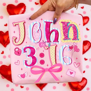 May include: A pink t-shirt featuring the text "John 3:16" in a decorative style. The design incorporates hearts, a bow, and other romantic motifs. The shirt is held up by a hand.