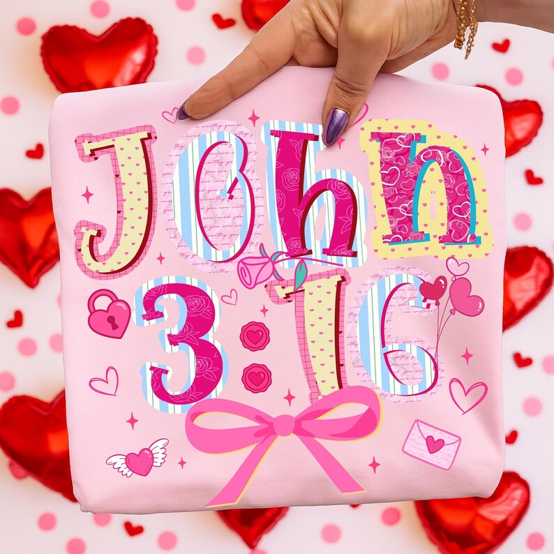 May include: A pink t-shirt featuring the text "John 3:16" in a decorative style. The design incorporates hearts, a bow, and other romantic motifs. The shirt is held up by a hand.