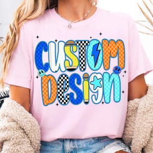 May include: Pink t-shirt with the words "CUSTOM DESIGN" in a colorful, playful font. The design incorporates elements like a lightning bolt, star, pencil, and checkerboard patterns. The shirt has a crew neck.