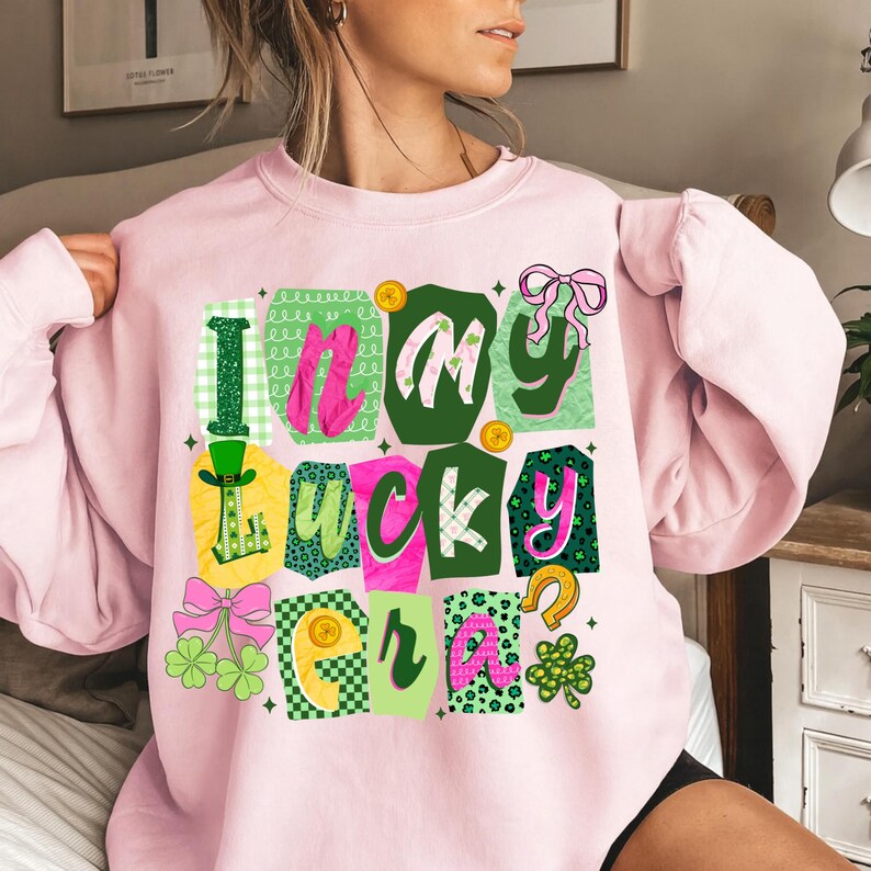 May include: A light pink sweatshirt with the text "I'm in my lucky era" in a colorful, patchwork design. The design includes shamrocks, a horseshoe, and a leprechaun hat, perfect for St. Patrick's Day.
