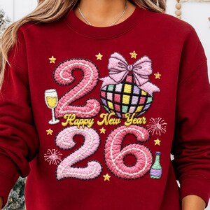 May include: A maroon sweatshirt with embroidered New Year's design. The design includes the numbers "26" in pink, a disco ball, champagne glass, bow, stars, and the text "Happy New Year".