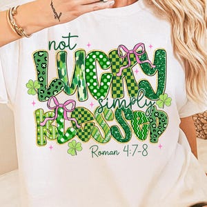 May include: White t-shirt with a St. Patrick's Day graphic. The design features the words "not LUCKY simply BLESSED" in green with various patterns, pink bows, and shamrocks. The text "Roman 4:7-8" is also included.