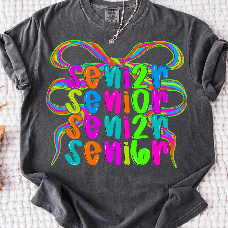 May include: Dark gray t-shirt with the word "Senior" in colorful, layered text. The word is in orange, blue, green, and pink, with a rainbow-colored bow design above. A silver necklace is visible.