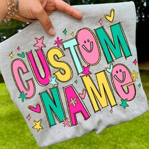 May include: A gray sweatshirt with the words "CUSTOM NAME" in a colorful, playful font. The letters are outlined in black and gold, with pink, yellow, and teal colors. The design includes stars, hearts, and smiley faces.