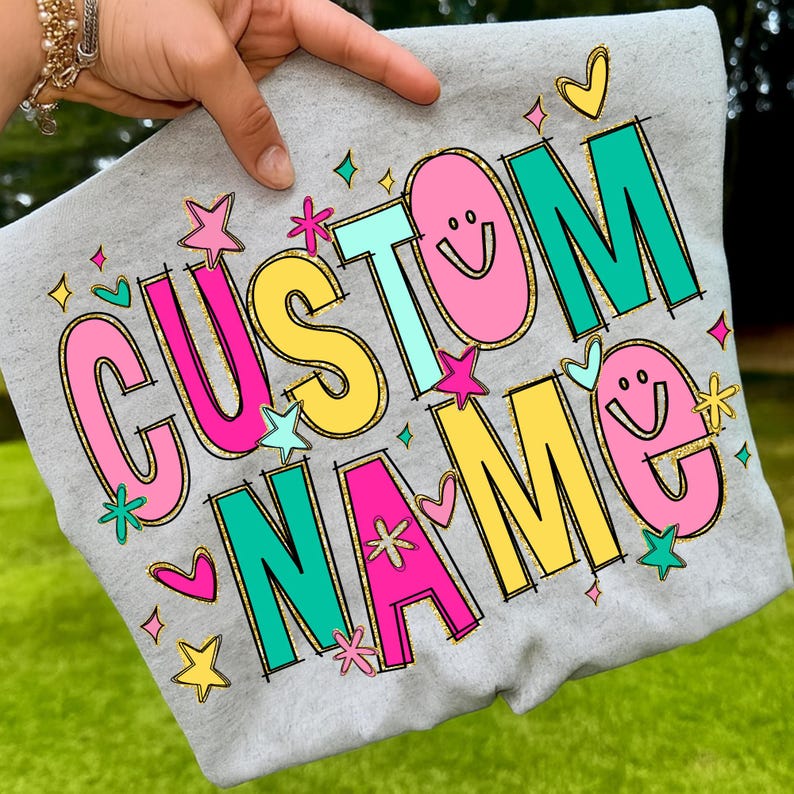 May include: A gray sweatshirt with the words "CUSTOM NAME" in a colorful, playful font. The letters are outlined in black and gold, with pink, yellow, and teal colors. The design includes stars, hearts, and smiley faces.