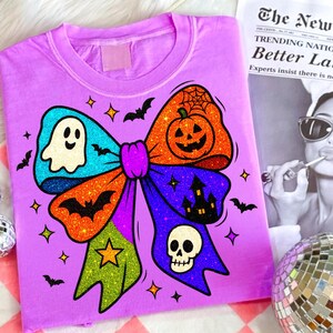 May include: A lilac-colored t-shirt featuring a large, glittery bow design with Halloween-themed elements. The bow includes a ghost, pumpkin, bats, a haunted house, and a skull. The shirt is decorated with stars and bats.