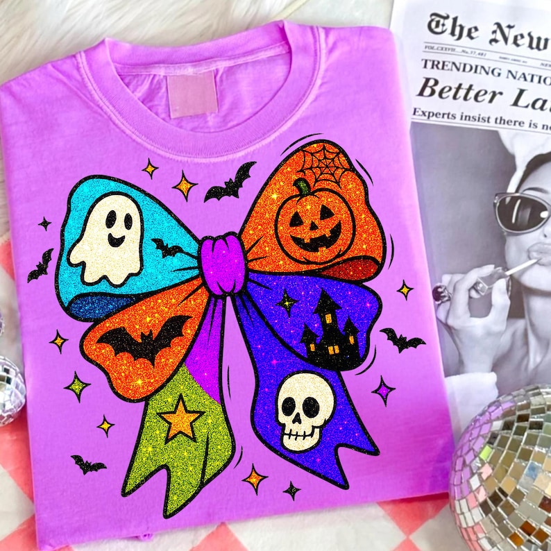 May include: A lilac-colored t-shirt featuring a large, glittery bow design with Halloween-themed elements. The bow includes a ghost, pumpkin, bats, a haunted house, and a skull. The shirt is decorated with stars and bats.