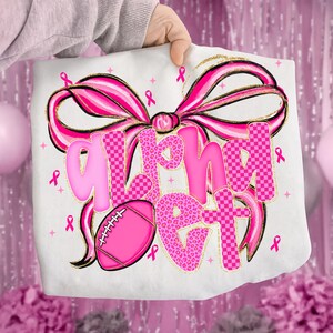 May include: White t-shirt with a large pink bow and the words "Alpha Get" in pink. The letters have a mix of leopard print and checkered patterns. A pink football and pink ribbons are also featured.