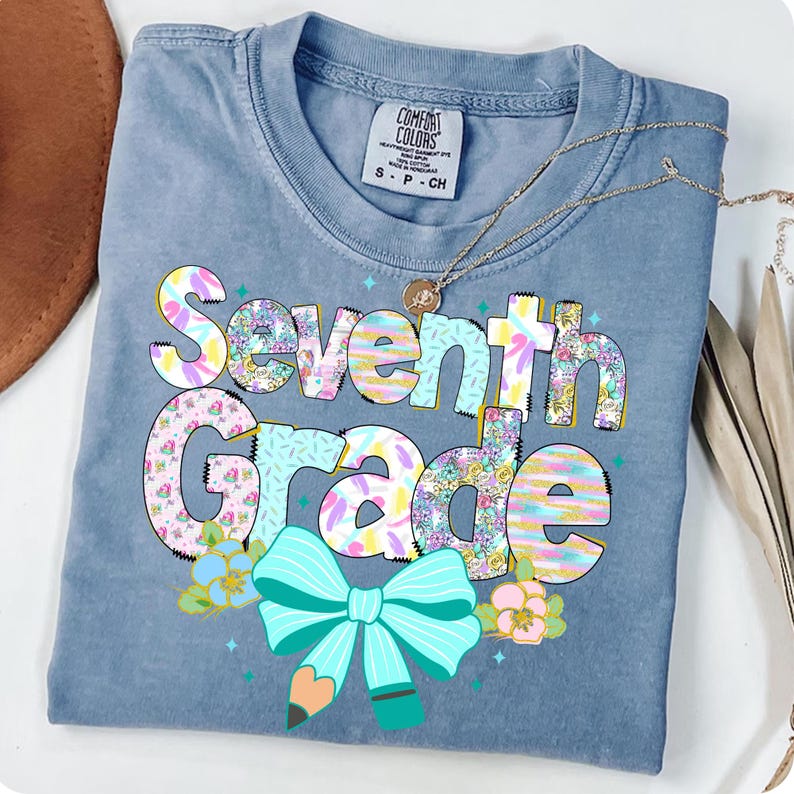 Op de afbeelding: Een blauw Comfort Colors T-shirt met de woorden "Seventh Grade" in een kleurrijk, bloemig ontwerp. Het ontwerp bevat een turquoise strik en een potlood. Een gouden ketting is over het shirt gedrapeerd.